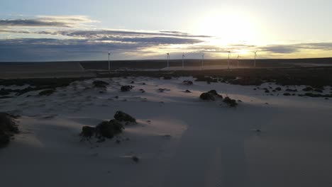 Antena-De-Drones-Cerca-De-Molinos-De-Viento-Que-Se-Desplazan-Hacia-La-Derecha-Durante-Un-Amanecer-Con-Dunas-De-Arena