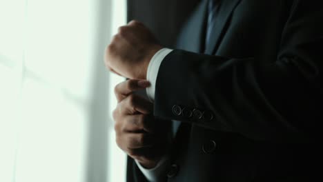 confident businessman in classic blue suit buttoning or adjust cufflinks near window in hotel room at the morning. handsome man wearing a nice suit on wedding day.