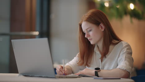 cheerful pretty cute nice red-haired girl girlfriend having been employed to job as executive smiling toothily sitting at desktop with laptop noting down important information