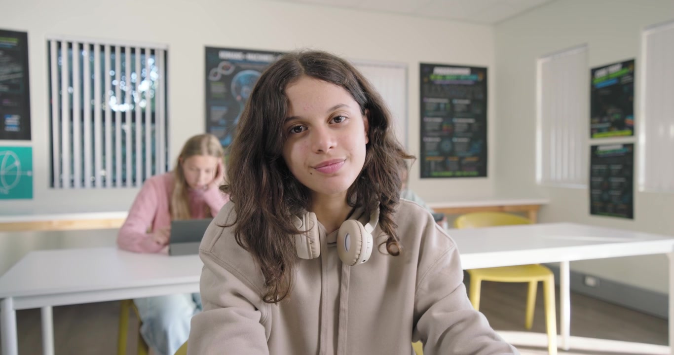 Weibliche Teenager-Klassenkameradinnen im Unterricht, wobei die vordere Teenagerin ein Tablet hält und die Kamera heranfährt, was ein Lächeln hervorruft.