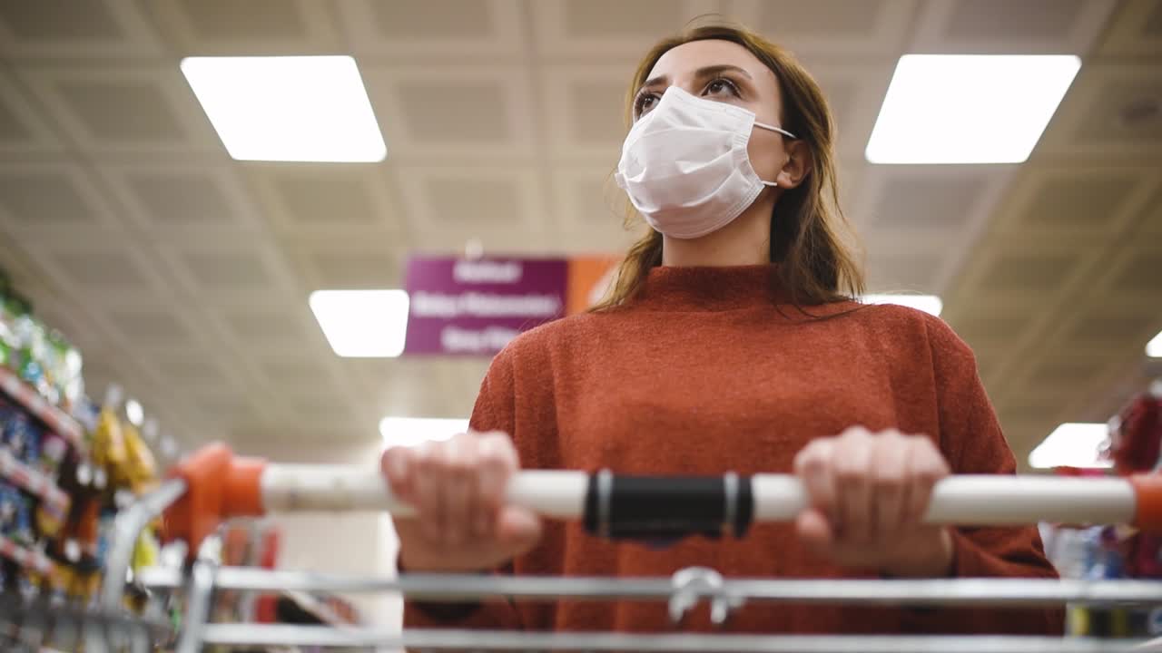 Beautiful woman wearing protective disposable medical mask and fashionable clothes uses shopping cart while shopping in supermarket