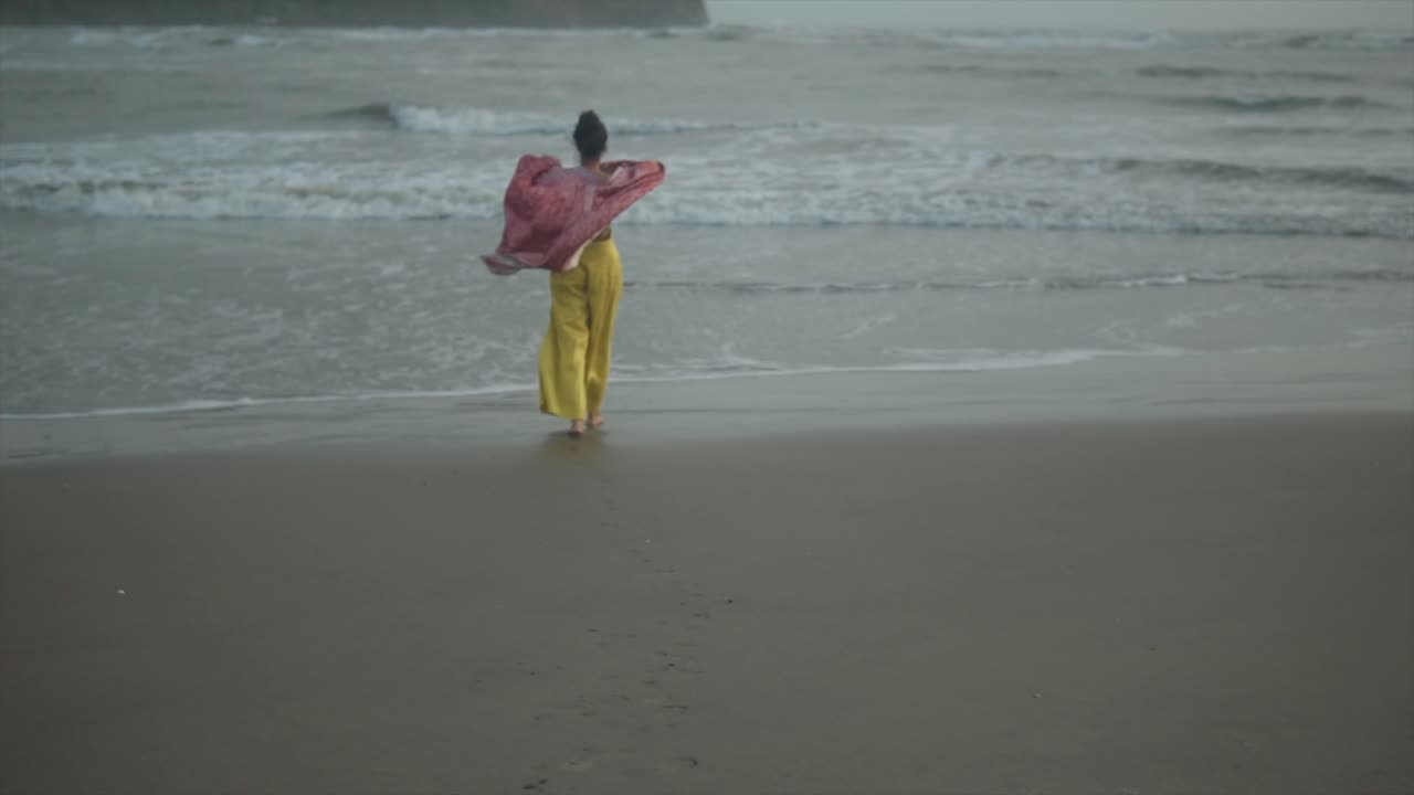 A slow-motion landscape view of a woman wearing yellow pants and holding a red scarf walking toward the sea