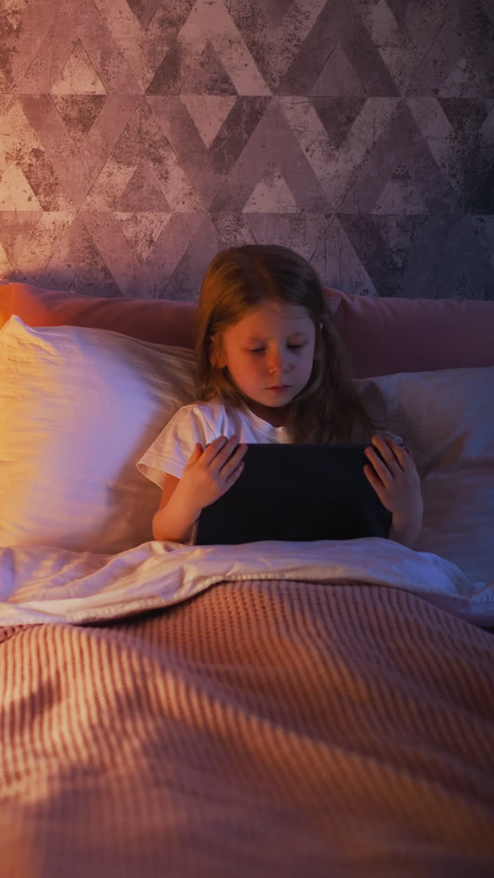 Blonde little girl with long hair uses tablet lying in comfortable bed with soft pillows and plaid near toy wigwam with glowing night lamp upper view slow motion