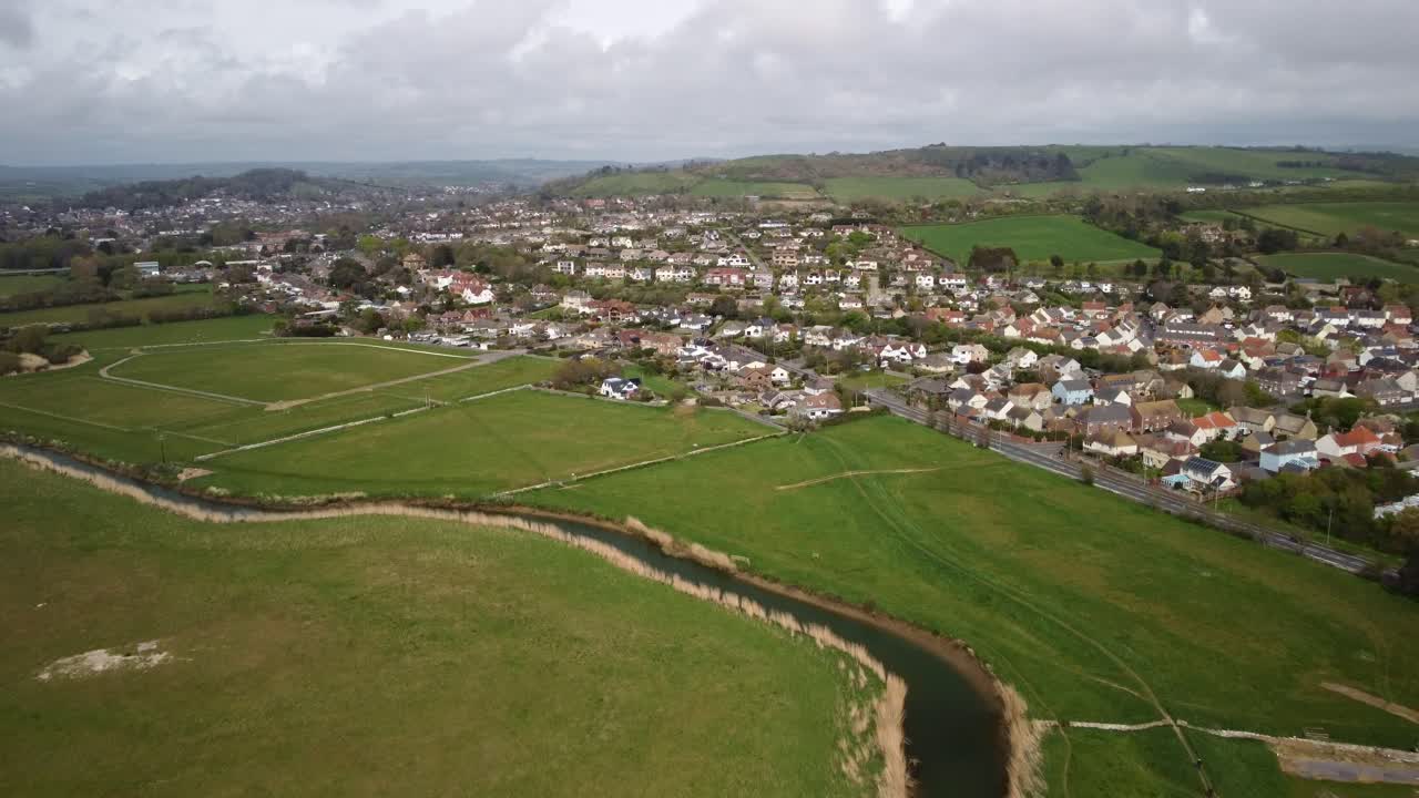 Drone video flying high over a river with fields either side and the a town, West Bay in Dorset behind. Beyond the homes are hills and a sky full of clouds. Green fields wide view forward movement.