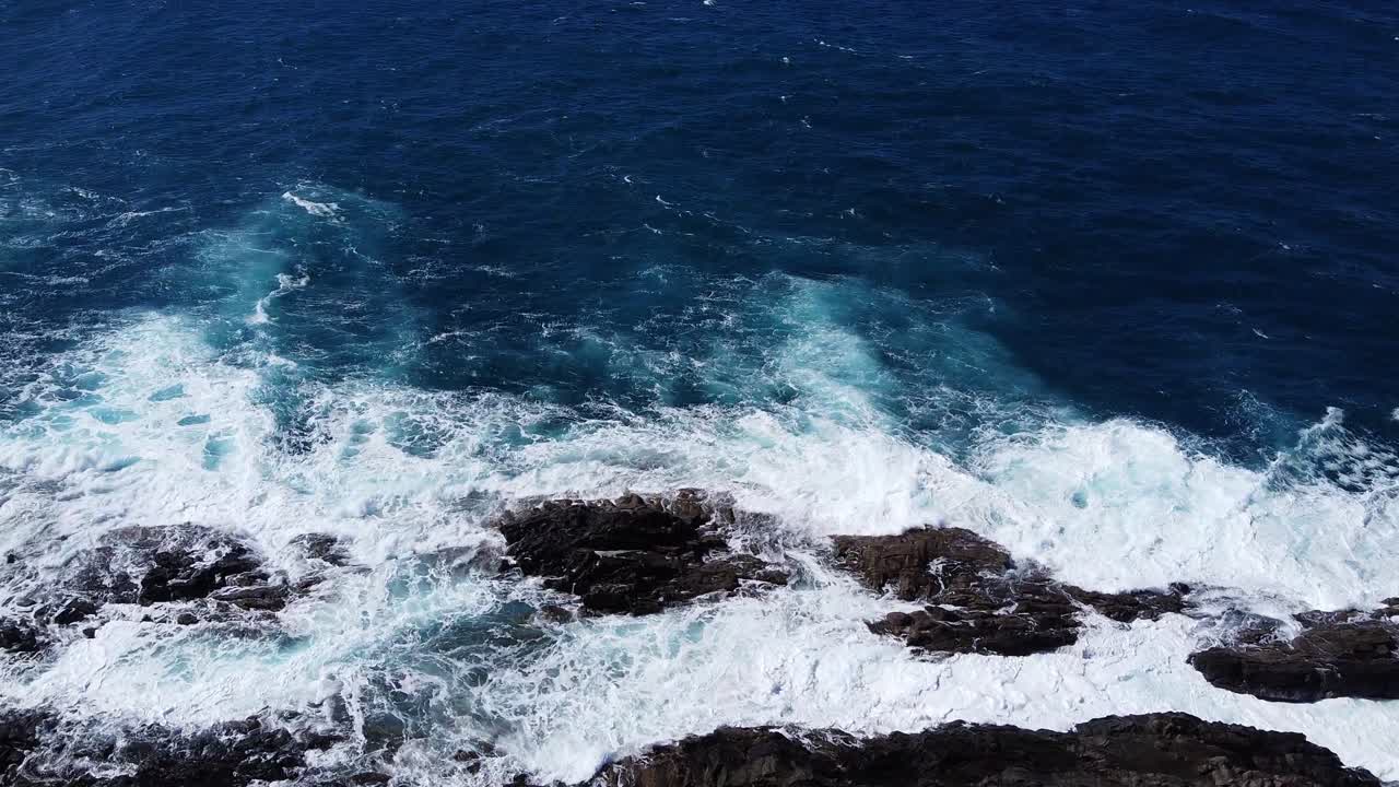 Deep blue waves of the ocean beat against the brown stones of the shore. Massive white foam and spray from. close up, midday son