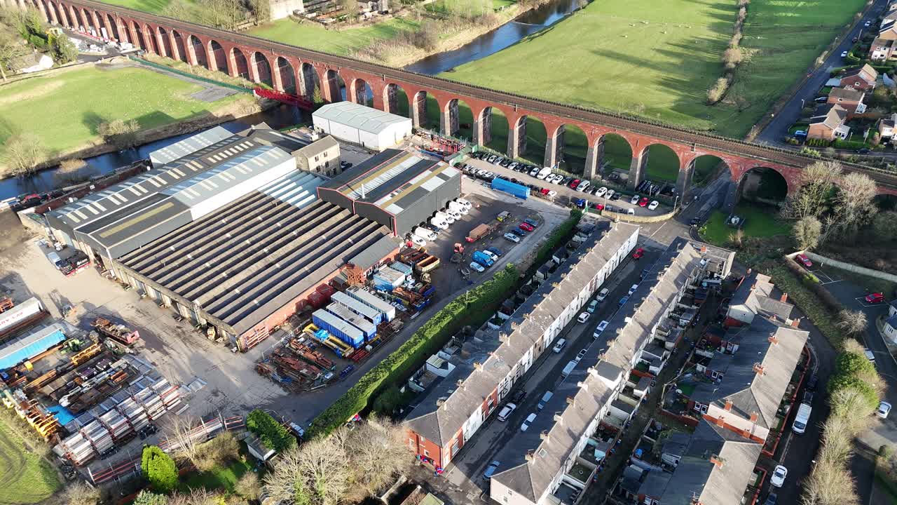 Drone video revealing a charming UK townscape with a historic red-brick viaduct. Sunlit factories, homes, and lush fields create a serene, picturesque setting from above