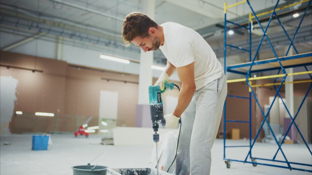 A construction worker skillfully mixes paint with a power drill, preparing for a renovation project in an industrial setting with scaffolding and tools nearby