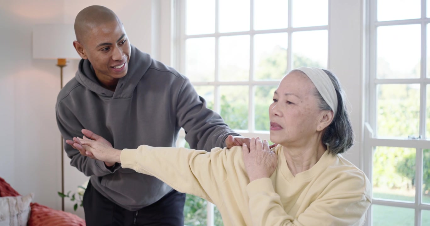 Senior Asian woman and caregiver at home, taking hand guiding arm on exercise ball for rehab