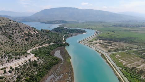 drone view in albania flying over wide blue river and green landscape next to the sea and road with mountains on the back