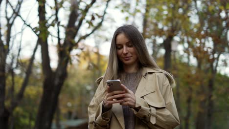 Lächelnde-Frau,-Die-Im-Herbstpark-Nachrichten-Liest-Oder-Vom-Telefon-Aus-Im-Internet-Surft,-Übersichtsaufnahmen