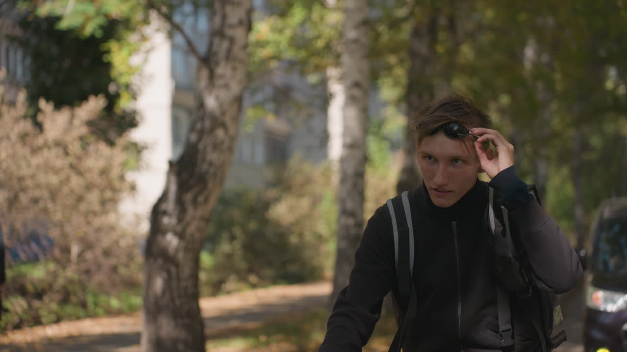 Uomo caucasico che si sistema gli occhiali da sole in bici, si ferma vicino a betulle e palazzi, ritratto di un pendolare o studente pensieroso, luce solare soffusa e a macchie e giacca casual, spallacci dello zaino