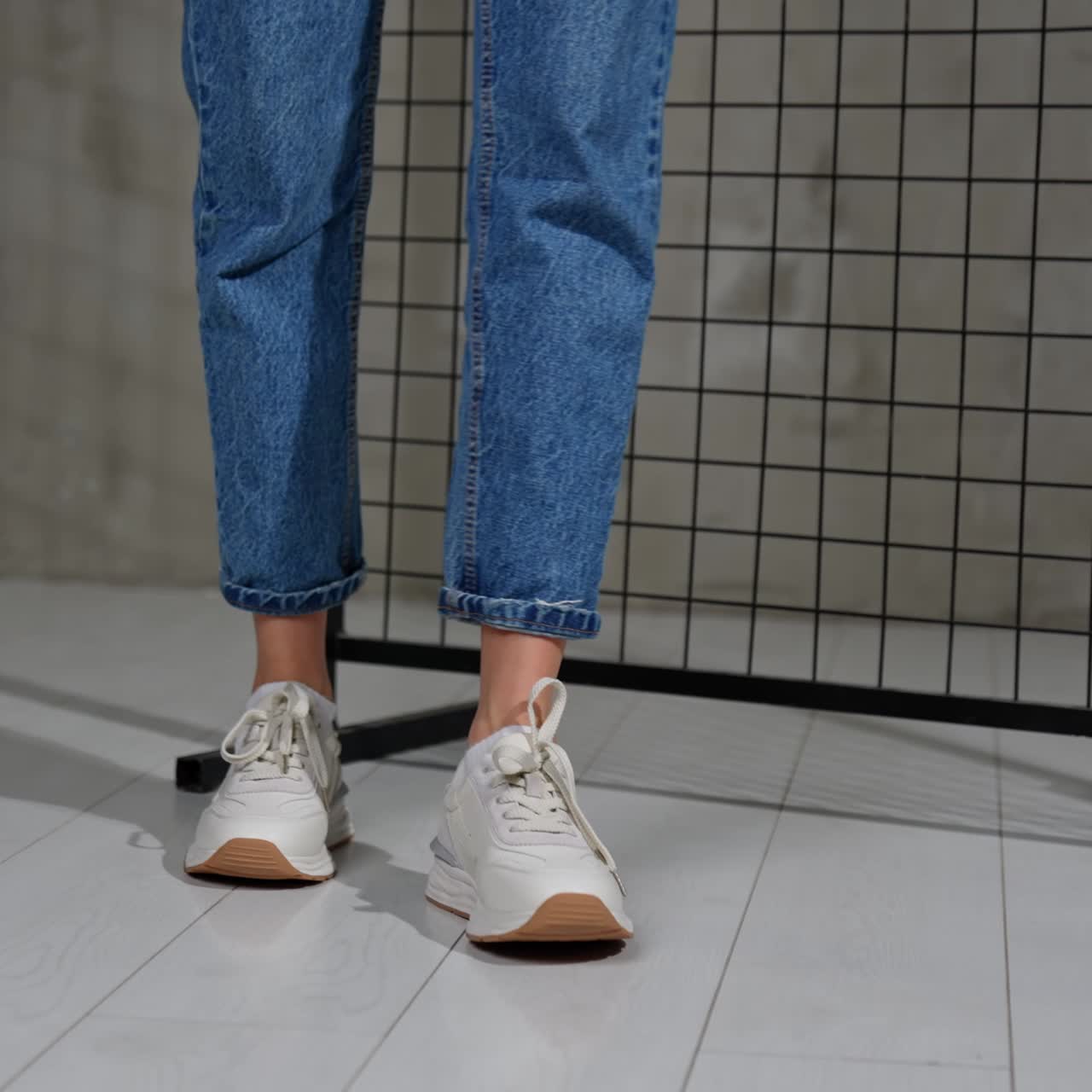 Girl turns around demonstrating the comfortable white sneakers. Female in blue jeans walks in stylish footwear. Black grate at backdrop