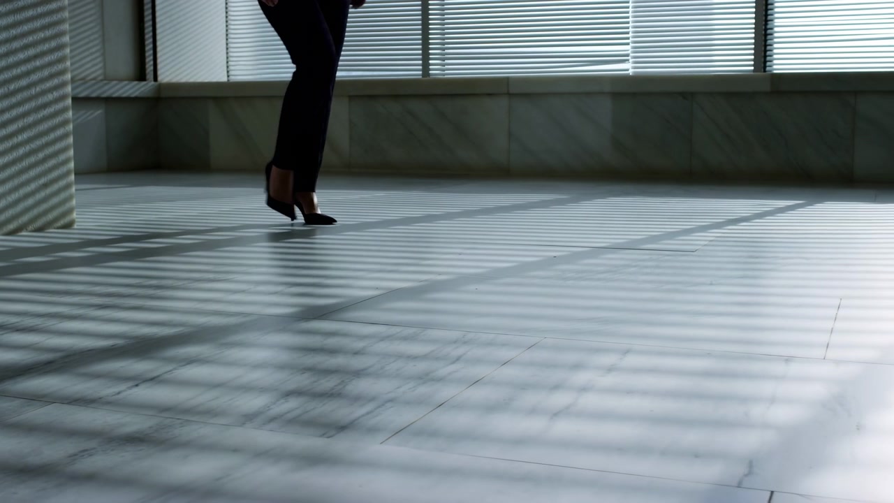 Bir iş kadını modern bir ofis koridorunda yürüyor. Resmi kıyafet ve topuklu ayakkabı giymiş, çizgili perdelerin oluşturduğu güneş ışığı alan bir zeminde adımlıyor.