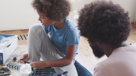 Over-shoulder-view-of-pre-teen-boy-sitting-on-the-floor-in-the-living-room-with-his-father,-using-tools-to-assemble-a-kit-toy,-close-up