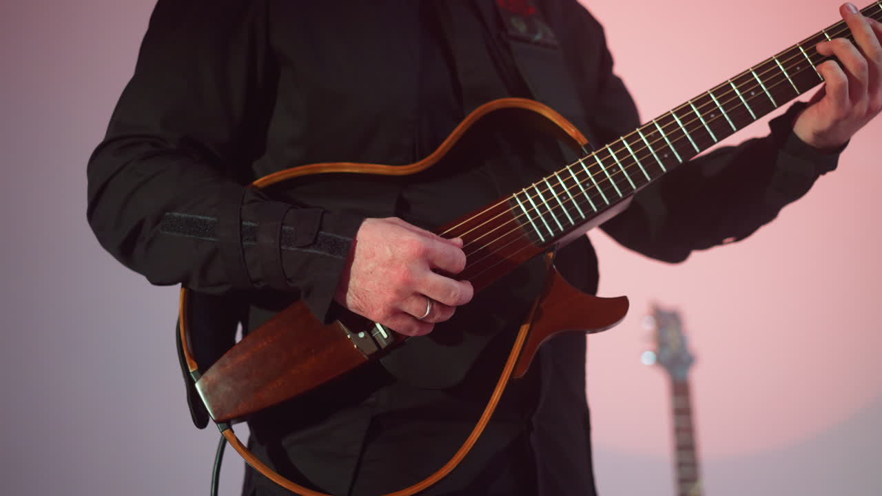 Guitarist's focused hand playing a red guitar against a softly blurred grey background, with another guitar faintly visible in the backdrop
