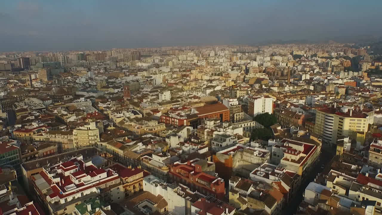 Ripresa aerea che avanza sopra i tetti di Málaga sotto una luce diurna soffusa con un cielo leggermente velato e un tessuto urbano denso