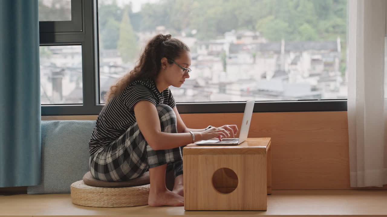 Female typing indoors. Young lady studying with laptop by window. Female using her computer next to city view window. Young woman focused on her notes and typing from her hotel room window
