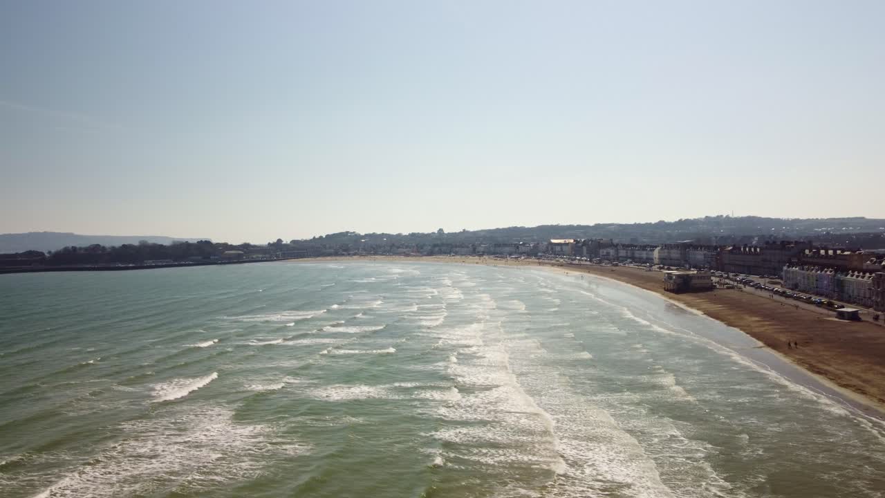 Aerial video moving over the water from Weymouth Beach, Dorset. The footage starts low over the beach and moves out to the left while raising. Show the bay and surrounding buildings, shops and homes.
