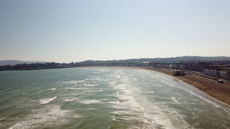 aerial video moving over the water from weymouth beach, dorset. the footage starts low over the beach and moves out to the left while raising. show the bay and surrounding buildings, shops and homes.