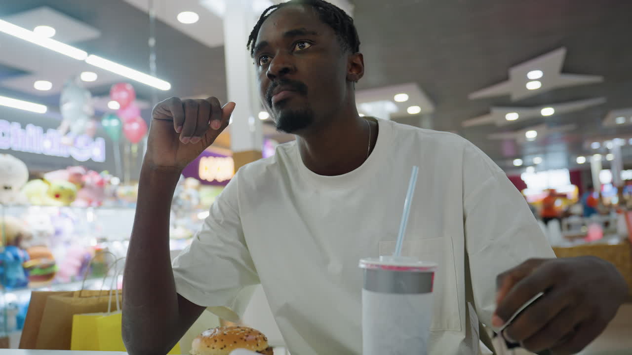 Man in white shirt seated in modern fast food restaurant drinks from disposable cup with straw while burger sits on tray in front of him amid colorful toy shop backdrop and blurred crowd activity