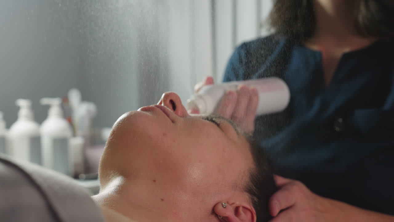 Beautician spraying fine hydrating mist over face of relaxed female client under bright spa light for instant skin refresh and soothing glow during advanced facial treatment session