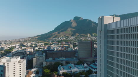 Forwards-fly-along-tall-building-in-city.-Elevated-footage-of-urban-borough-and-mountains-in-background.-Cape-Town,-South-Africa