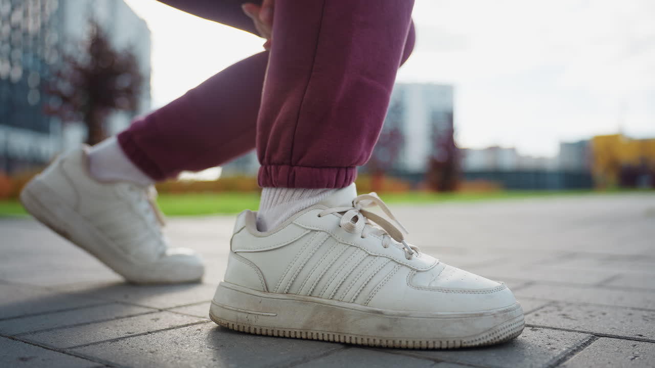 Strength trainer bends down to tie canvas sneaker lace on paved urban sidewalk outside modern apartments under warm calm morning light showcasing focused preparation for outdoor fitness routine