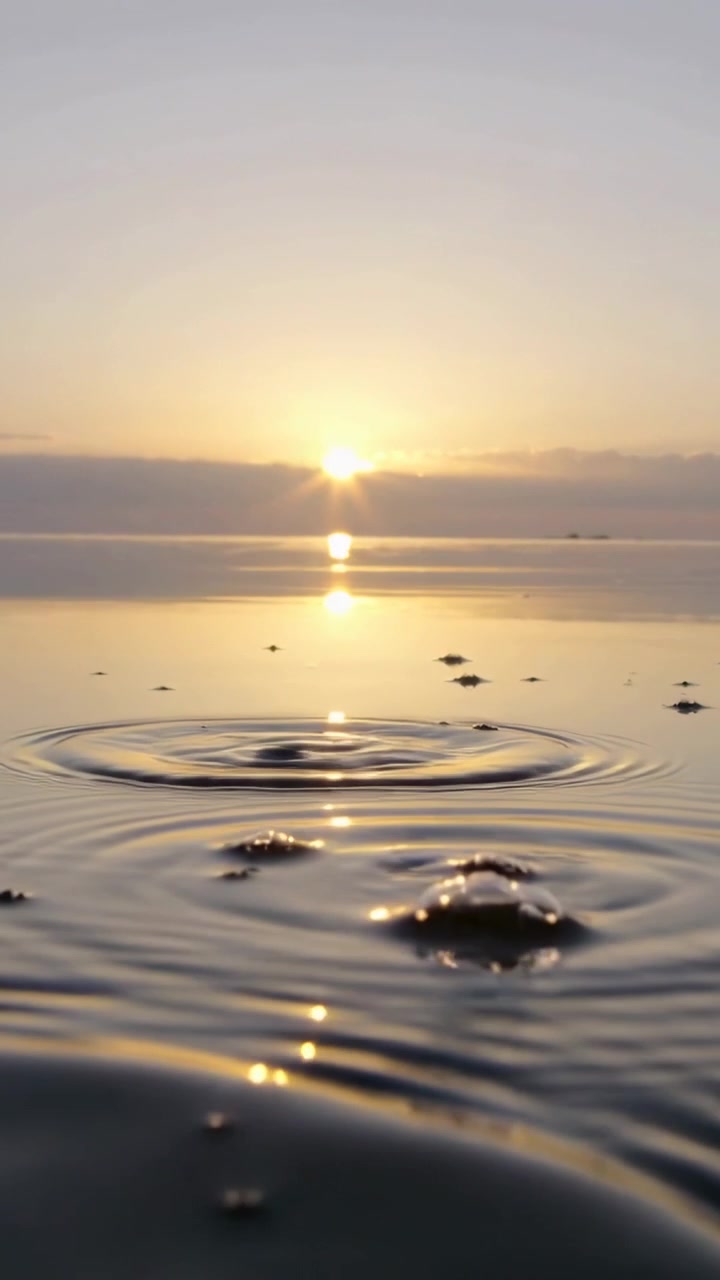 Video verticale: Increspature dopo lo scoppio di una goccia, il mare forma anelli all'alba con bagliore solare e chiazze.