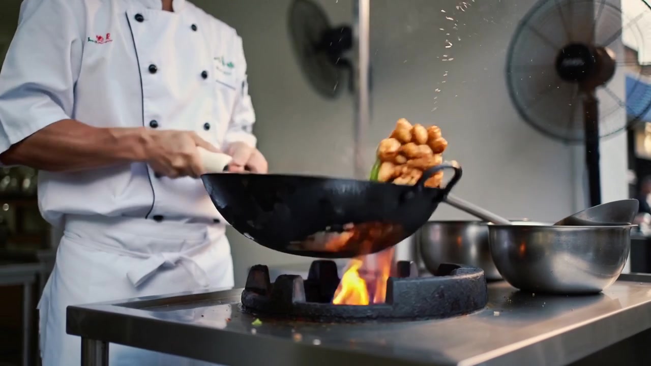 Uno chef con una giacca bianca solleva e fa saltare il wok su una fiamma viva, lanciando le verdure per scottarle.