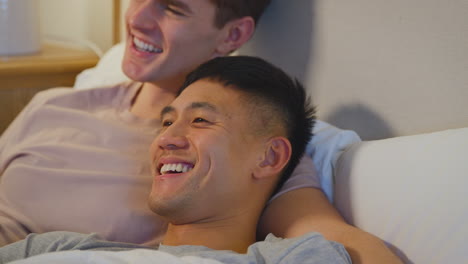 close up of loving same sex male couple lying in bed at home watching tv together