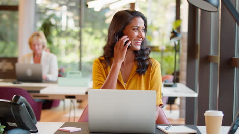 Mature-Businesswoman-Working-On-Laptop-At-Desk-In-Office-Taking-Call-On-Mobile-Phone