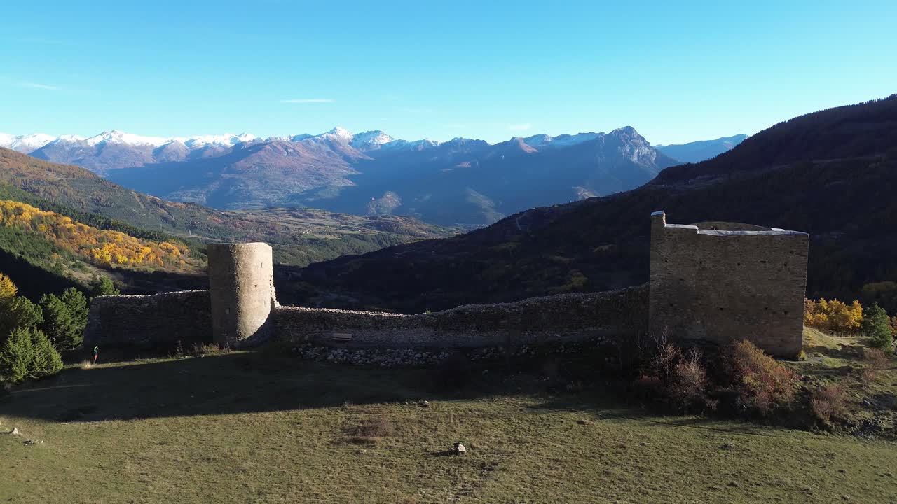 Drone captures the ruins of Fort de Crévoux with snowy alpine peaks in the distance. Colorful autumn trees and clear November sunlight highlight the dramatic landscape of the Hautes Alpes
