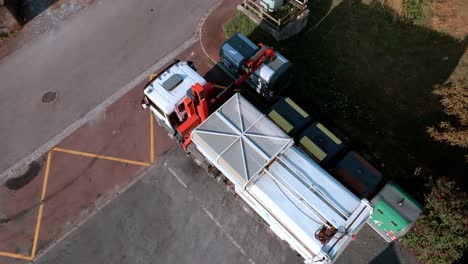 refuse truck is working with recycled glass from containers by the roadside, lifting up and unloading them in the back of the truck, top aerial drone view during sunshine day, glass recycling concept