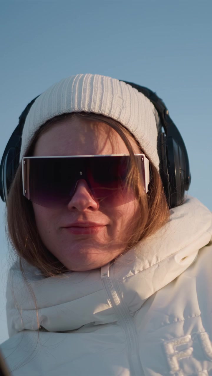 Woman wearing white jacket and headphones holds smartphone with stickers looking at screen. scene features soft natural light against outdoor background with blurred trees or landscape