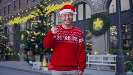 asian man in santa suit smiling and showing thumbs up gesture while standing in christmas night