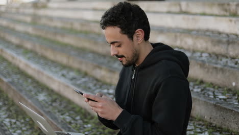 Young-Arabic-man-with-dark-curly-hair-and-beard-in-black-hoodie-sitting-on-stairs-outside