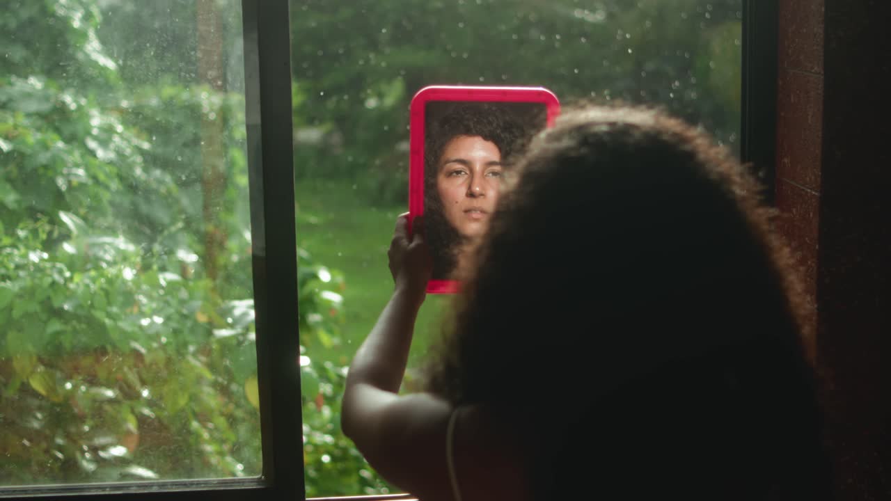 Camera pulls out from woman with voluminous curly hair standing by a window, holding a pink-framed mirror that reflects her face creating a layered visual of identity and perception
