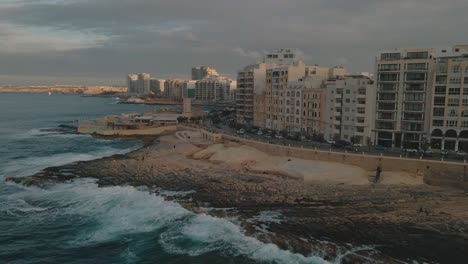 panning, aerial view of the seaside city of sliema, malta as waves crash onto the rocky beach