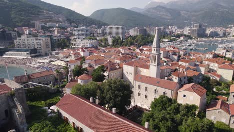 Church-of-Sveti-Ivan,-Budva,-Montenegro-with-scenic-coastal-town-backdrop---aerial