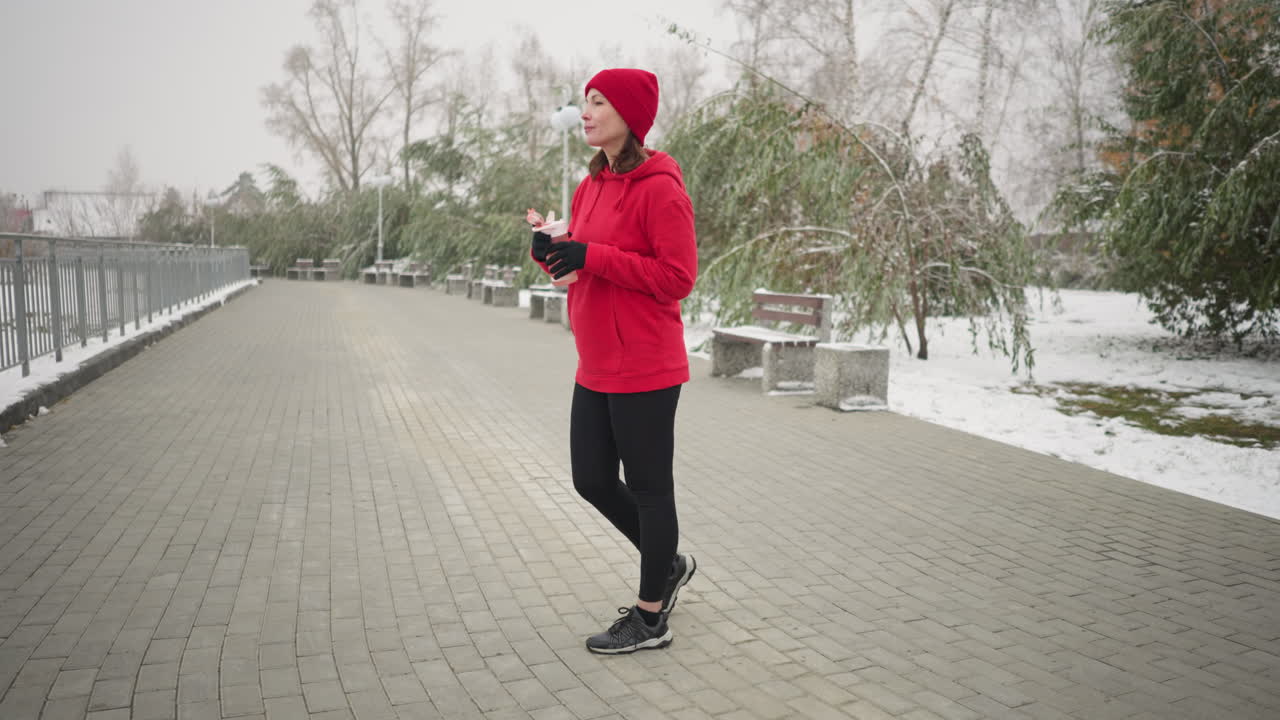 Lady in a pose opens water bottle and sips water from pink bottle outdoors in serene winter park setting with snow-covered ground, iron railing, benches, and trees in misty background