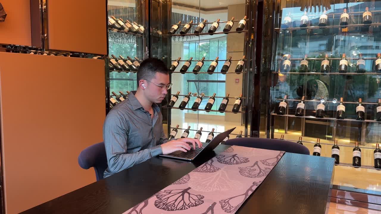 Homem asiático da geração milênio digitando em seu laptop, usando fones de ouvido e acenando com a cabeça enquanto trabalha dentro de uma casa luxuosa.