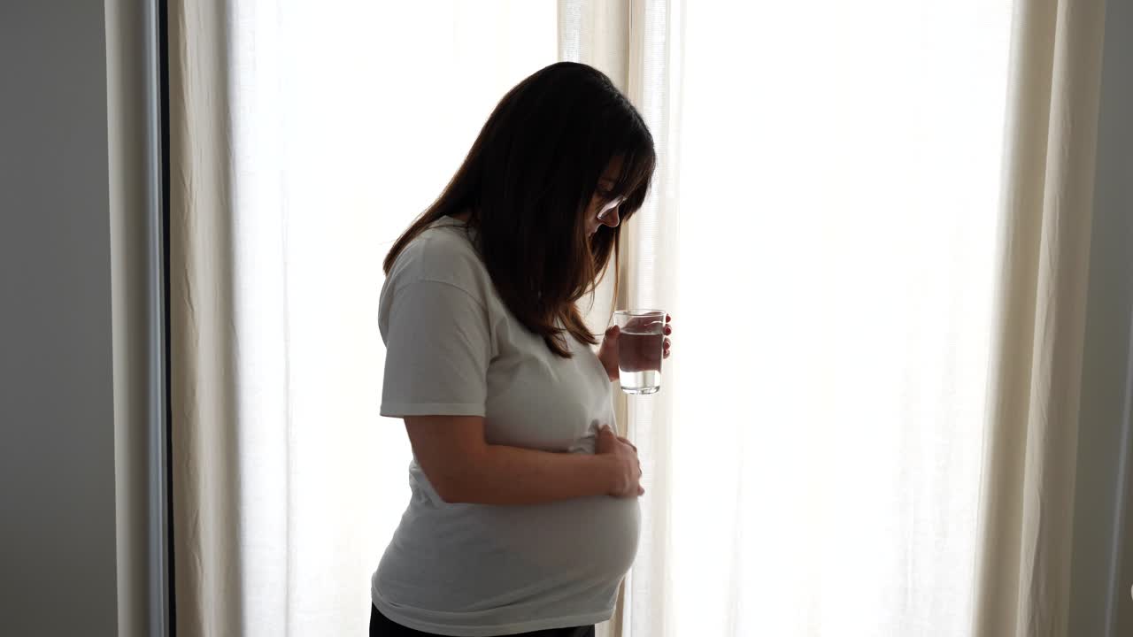 Pregnant woman drinks a glass of water standing by bright curtains in the bedroom
