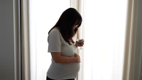 pregnant woman drinks a glass of water standing by bright curtains in the bedroom