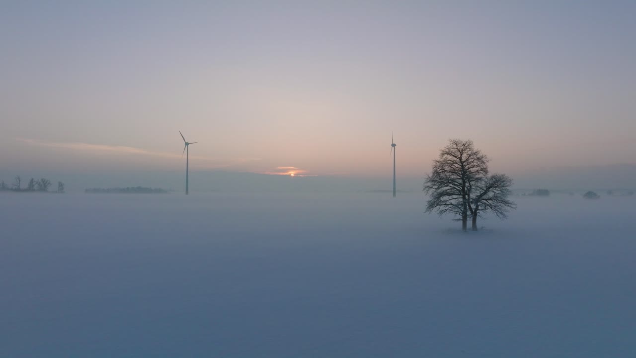 풍력 발전소에서 재생 에너지를 생성하는 풍력 터빈의 공중 전망, 안개로 가득 찬 눈으로 가득 찬 시골 풍경, 황금 시간의 빛이 있는 화창한 겨울 저녁, 넓은 드론 샷이 앞으로 낮게 움직입니다.