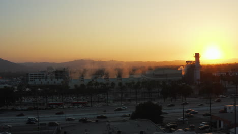 Antena:-Fábrica-Humeante-Con-Palmeras-Y-Carretera-Transitada-En-Burbank,-Los-ángeles,-California,-Puesta-De-Sol