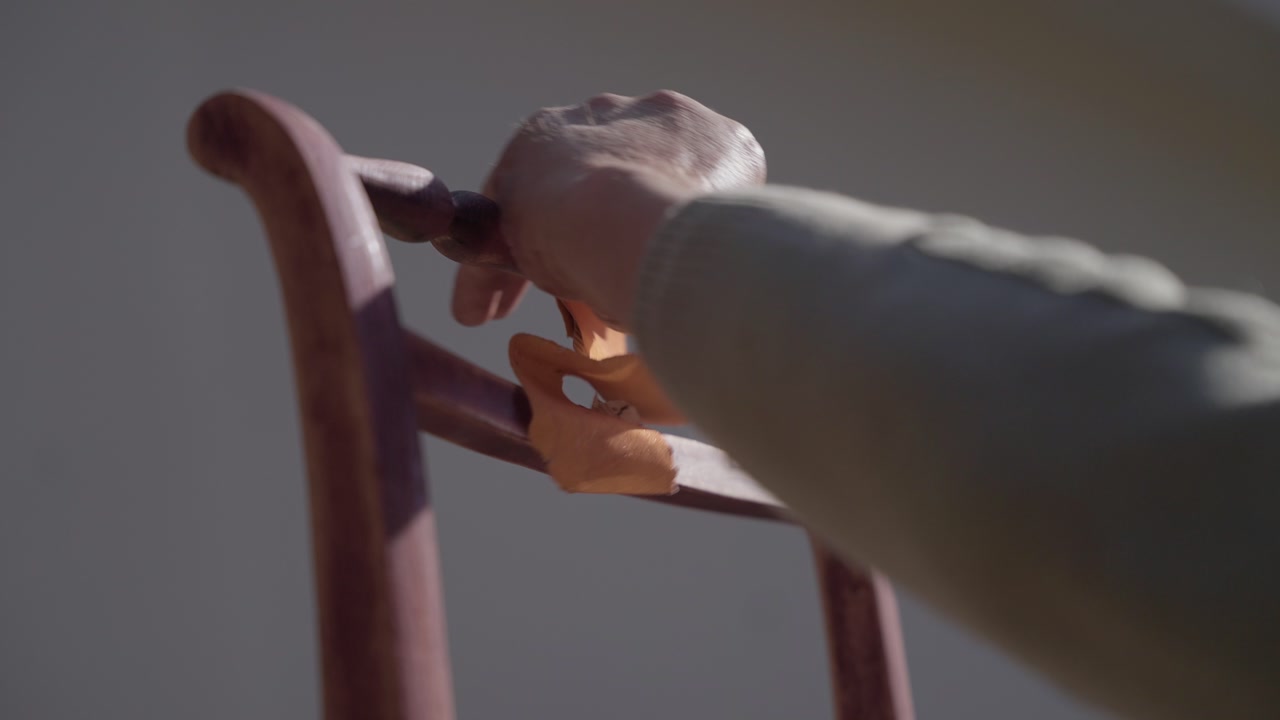 Closeup of hand rubbing sandpaper on curved wooden chair back during restoration outdoors