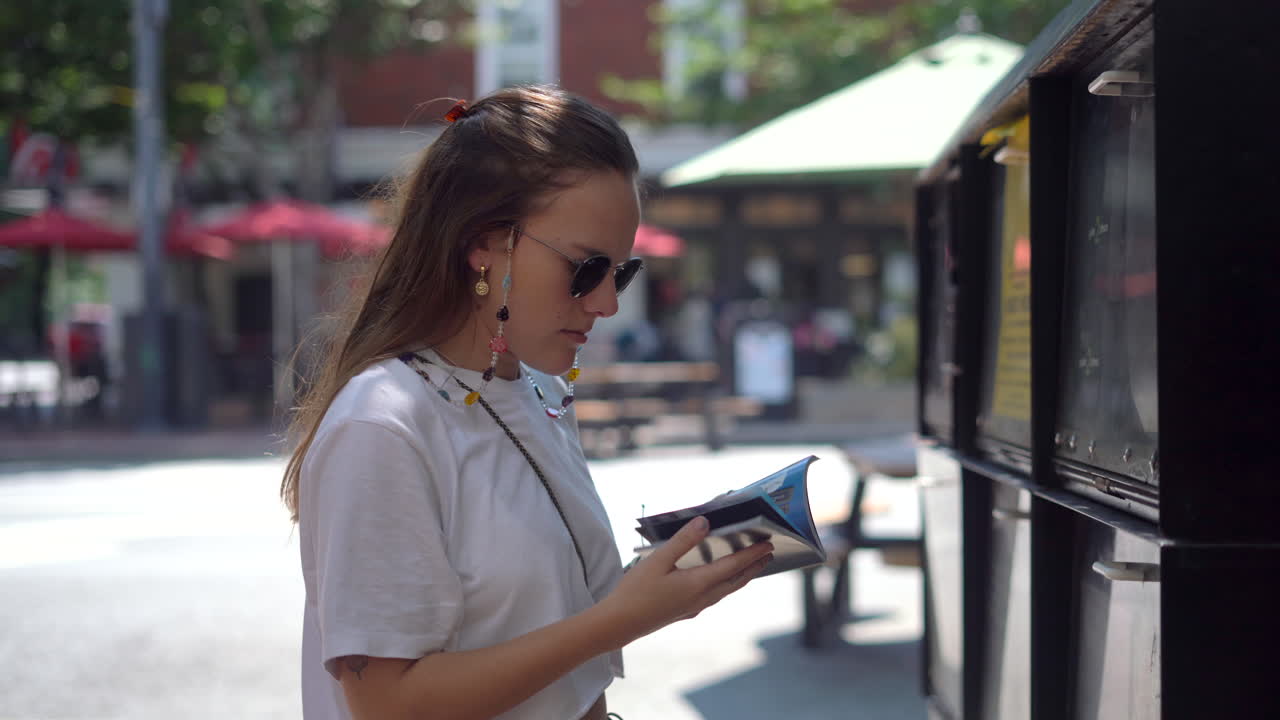 una joven toma una guía local gratuita o un calendario de eventos de un puesto de periódicos en un pequeño pueblo