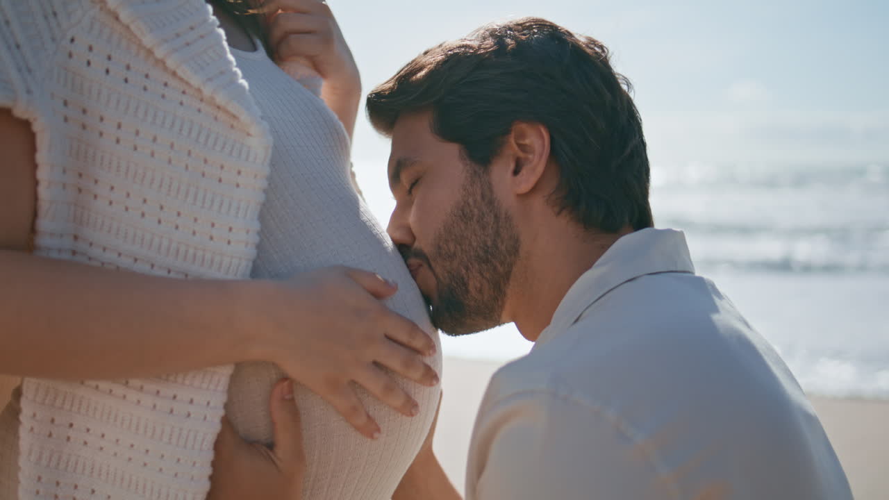 Future father caressing pregnant belly sitting near wife at sea coast close up.