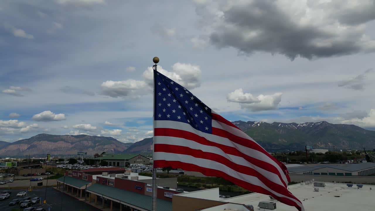 Bandiera americana USA che sventola al vento in una bella giornata di sole estiva con nuvole e cieli azzurri che si affaccia su montagne e una piccola città mentre il drone ruota rapidamente a sinistra - in 4K a 60fps