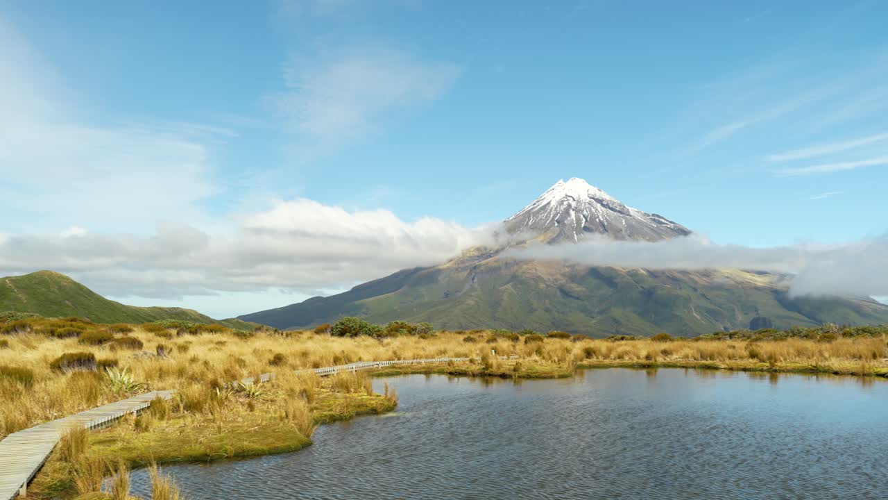 Yeni Zelanda'daki Pouakai Tarns'ta çekilmiş, yansıtıcı sulak alanı, altın rengi ot öbeğini ve alçak sürüklenen bulutların altında yükselen Taranaki Dağı'nı gösteren panoramik manzara çekimi.